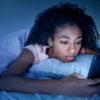 Mulher negra deitada na cama no escuro mexendo no celular com cara de preocupada.