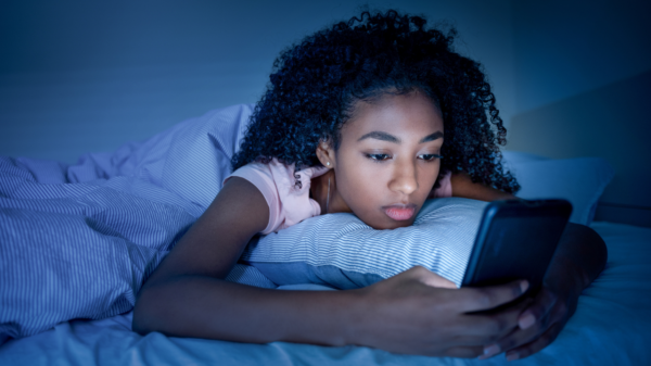 Mulher negra deitada na cama no escuro mexendo no celular com cara de preocupada.
