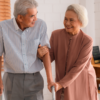 Um casal idoso. O homem está com andador e usa camisa e calça social. A mulher usa um vestido marrom. Ambos tem cabelo branco.