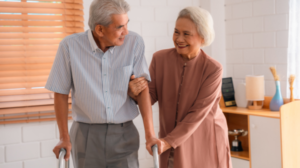 Um casal idoso. O homem está com andador e usa camisa e calça social. A mulher usa um vestido marrom. Ambos tem cabelo branco.