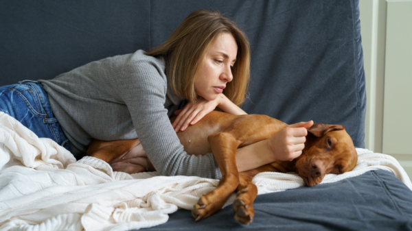 Mulher deitada no sofá abraçando um cachorro marrom que parece assustado ou desconfortável, oferecendo conforto e carinho ao pet.