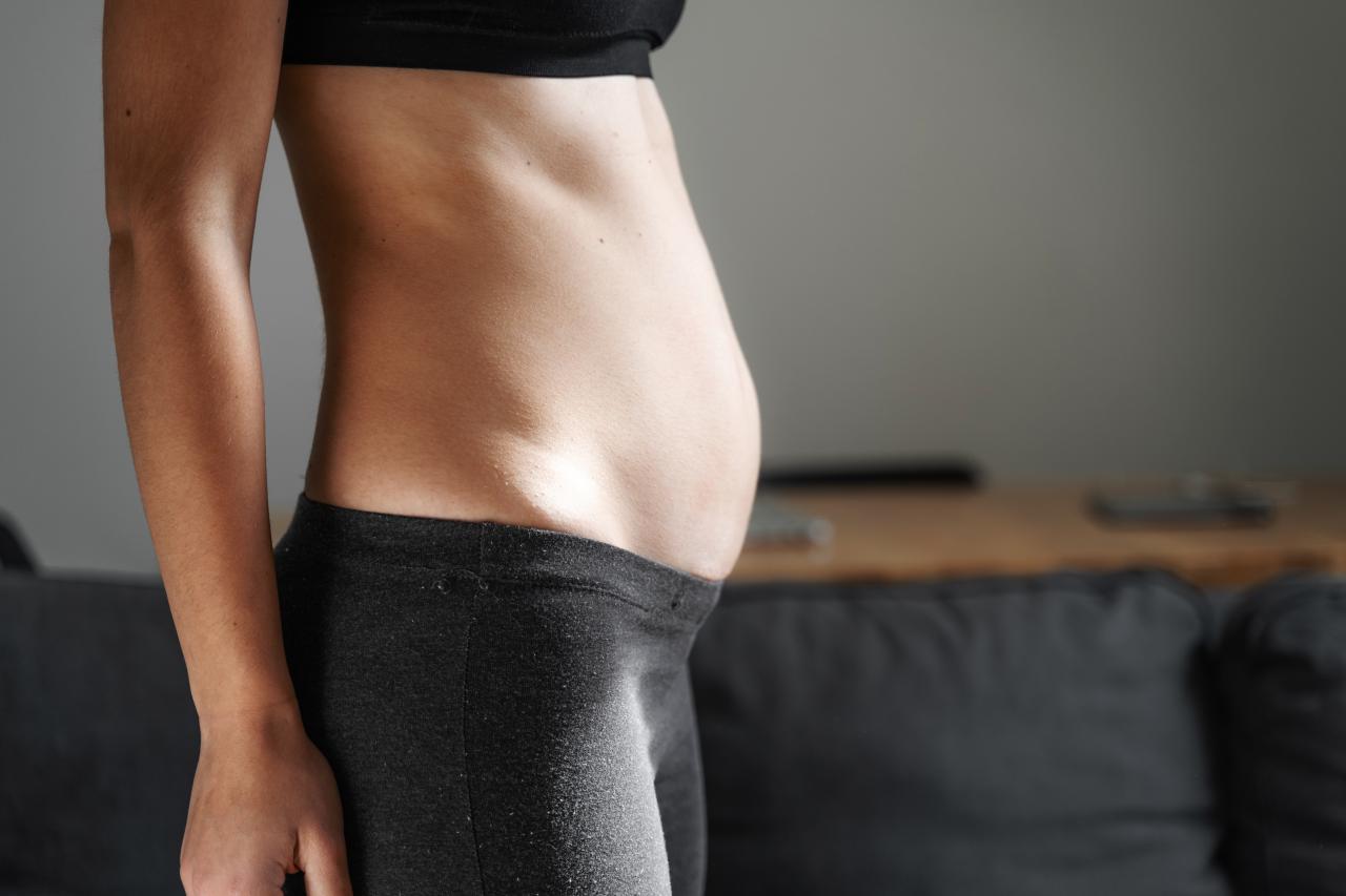 Pessoa em pé, vista de perfil, com destaque para a região abdominal levemente inchada, usando roupa esportiva, em ambiente interno.