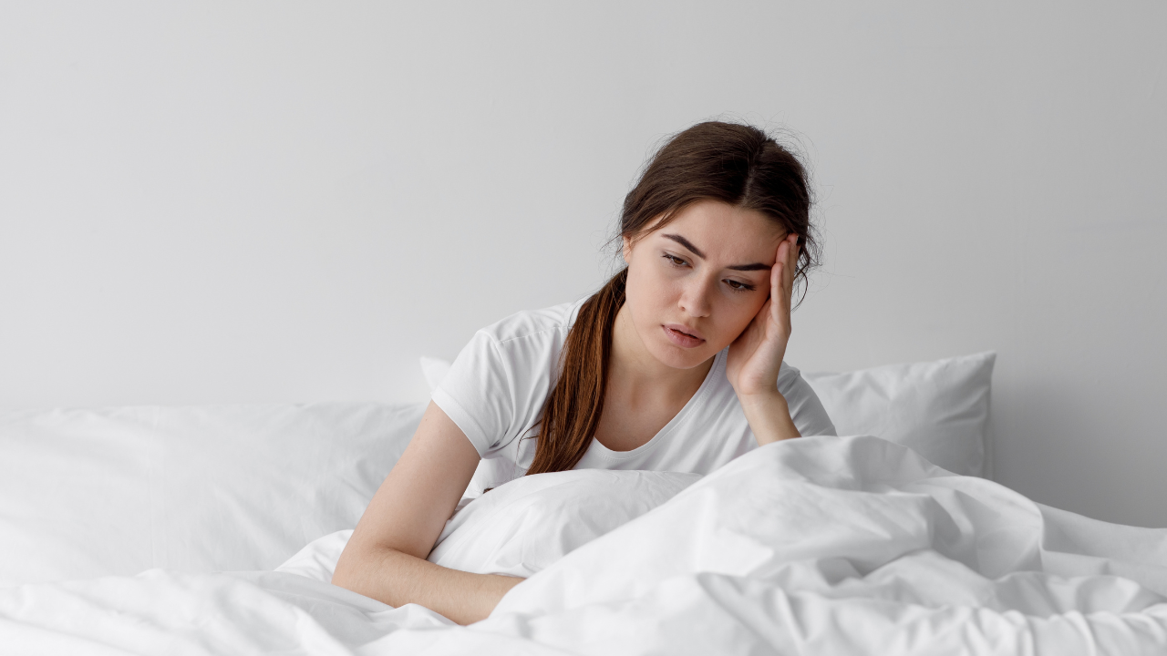 Mulher sentada na cama, coberta, com expressão de preocupação e dificuldade para dormir devido à ansiedade noturna.