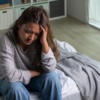Mulher sentada na cama, com a mão apoiada na cabeça e expressão de preocupação, em ambiente doméstico iluminado, representando ansiedade ou sofrimento emocional.