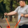 Homem branco, gordo, usando camiseta cinza, fazendo exercício em um parque.