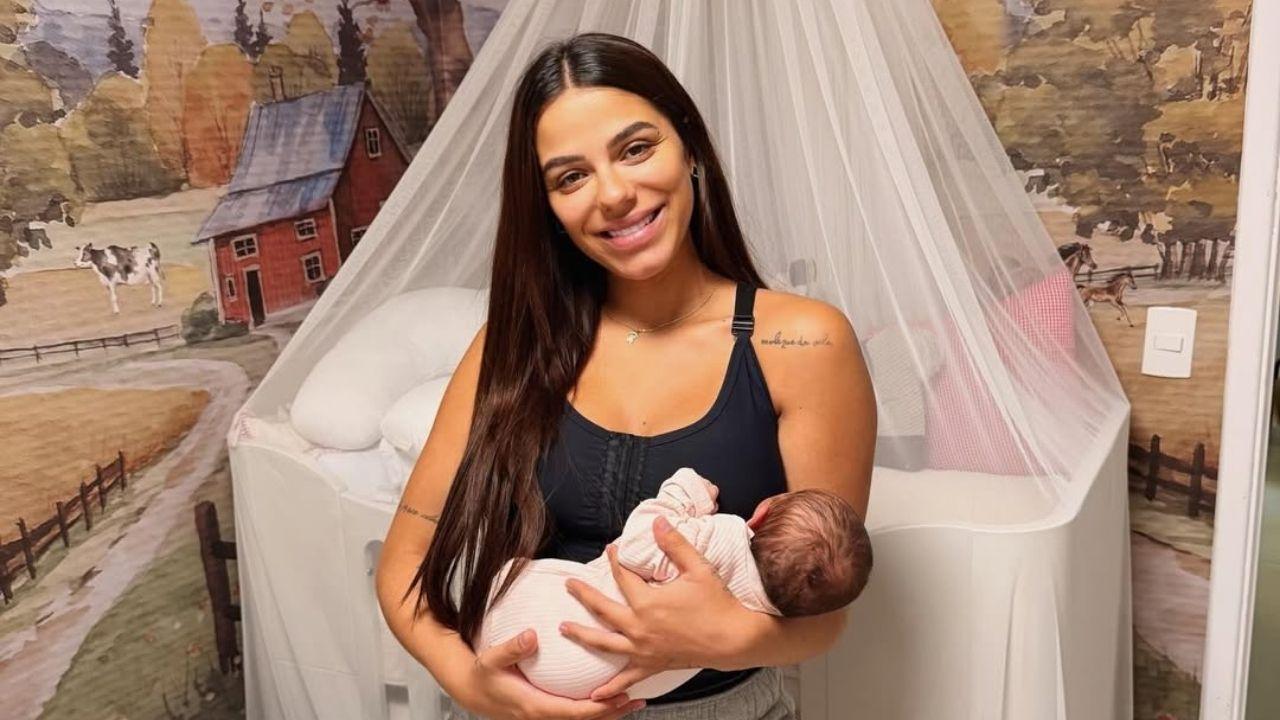 Mulher branca, cabelo liso solto, usando blusa preta e short branco segurando um bebê no quarto do bebê.