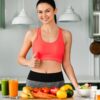 Mulher branca, magra, cabelo castanho preso fazendo sinal de joinha. Na mesa tem frutas, legumes e sucos.