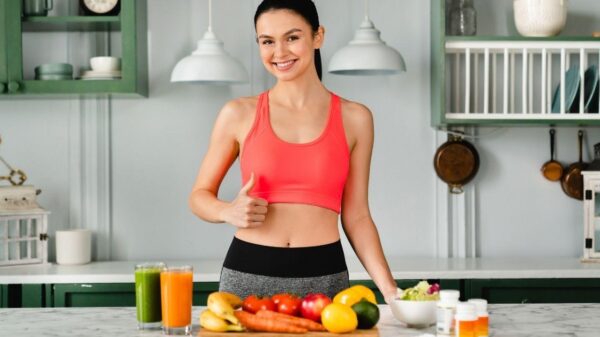 Mulher branca, magra, cabelo castanho preso fazendo sinal de joinha. Na mesa tem frutas, legumes e sucos.