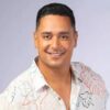 Xanddy Harmonia. Homem pardo, sorrindo, vestindo camiseta branca com flor em um fundo neutro.