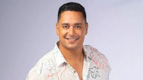 Xanddy Harmonia. Homem pardo, sorrindo, vestindo camiseta branca com flor em um fundo neutro.