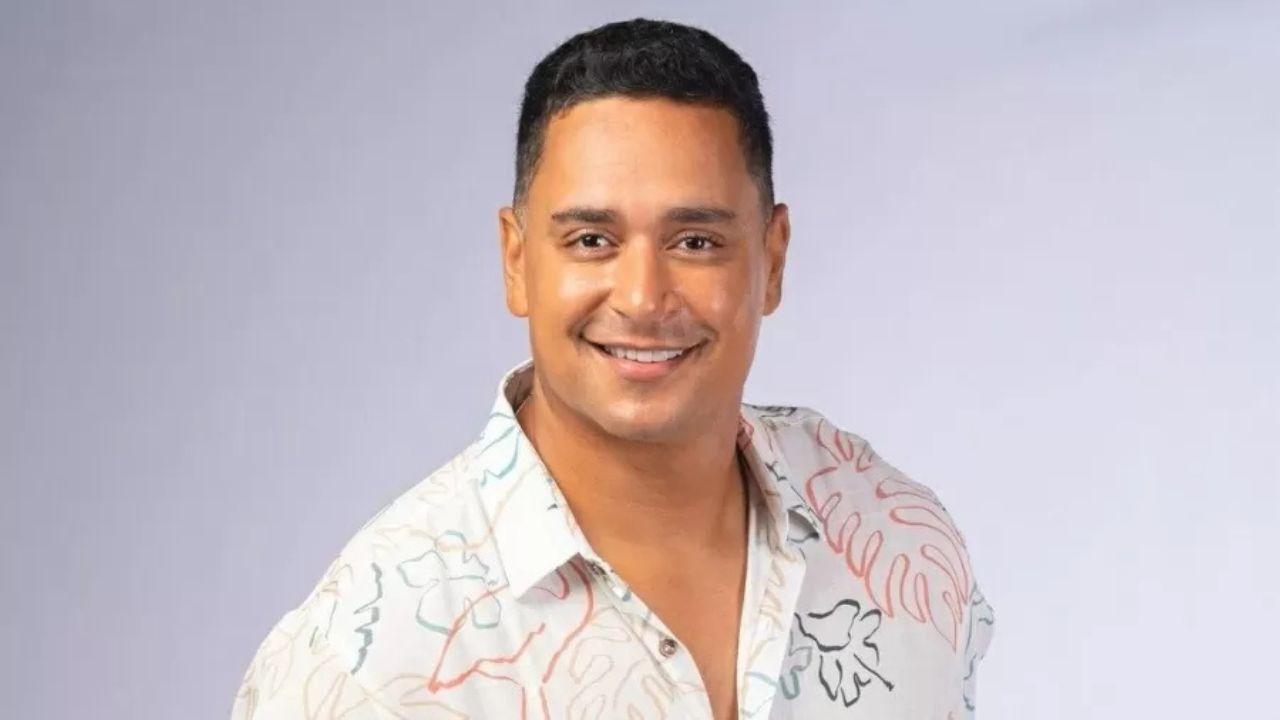 Xanddy Harmonia. Homem pardo, sorrindo, vestindo camiseta branca com flor em um fundo neutro.