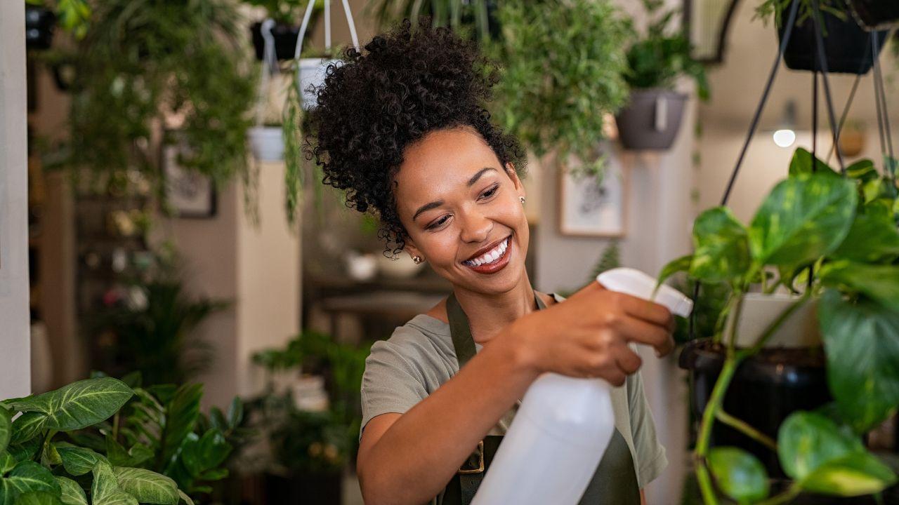 Mulher negra, cabelo cacheado preso em coque, usando avental e regando as plantas.