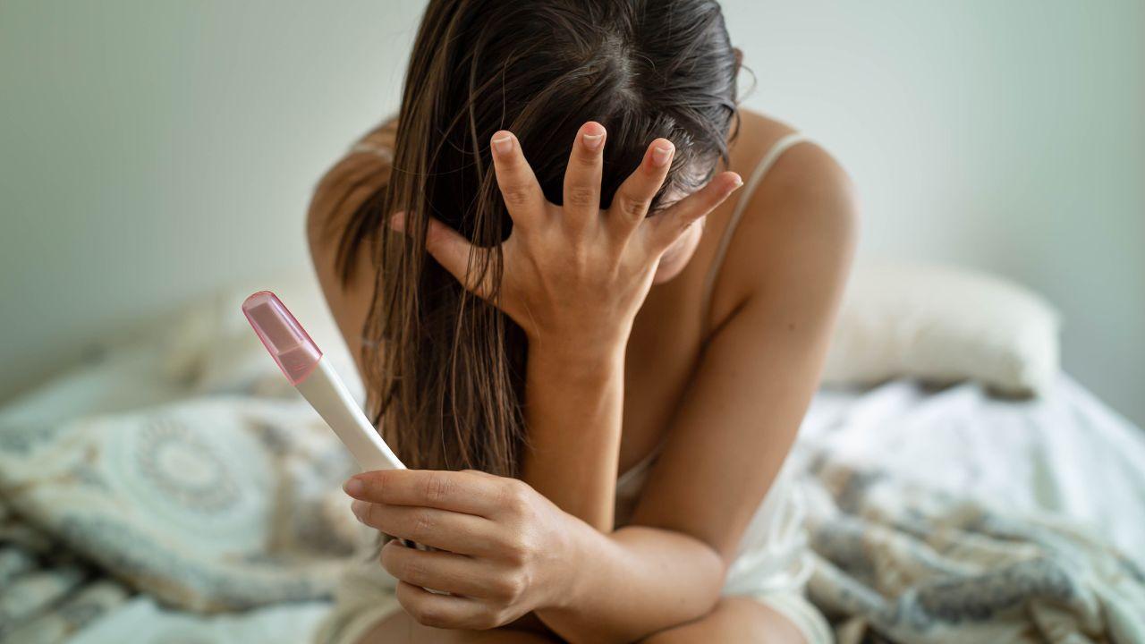 Mulher com a mão no rosto e segurando um teste de gravidez com semblante preocupado.