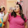 Imagem de uma mulher, de blusa rosa e tiara com lírios rosa, bebendo água no bloquinho
