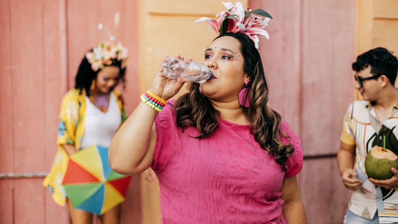 Imagem de uma mulher, de blusa rosa e tiara com lírios rosa, bebendo água no bloquinho