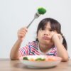 Imagem de uma menina olhando para um brócolis em um garfo. Há um prato com mais vegetais na frente dela