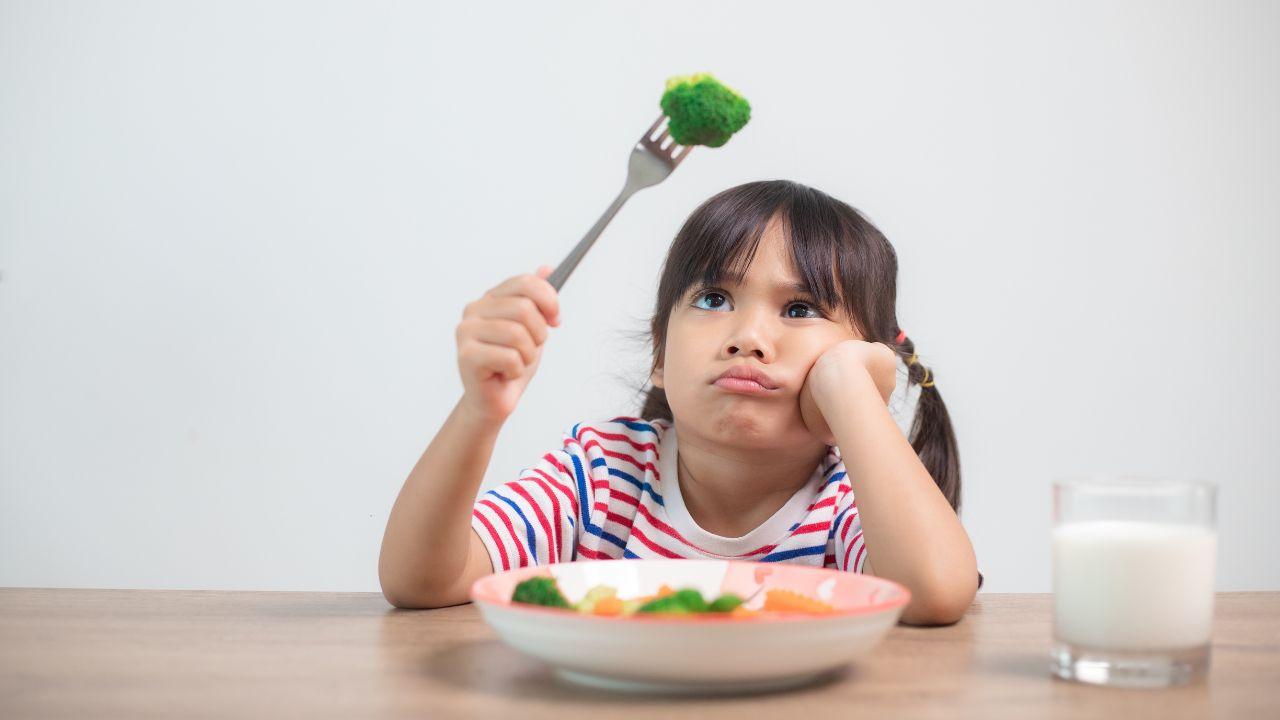 Imagem de uma menina olhando para um brócolis em um garfo. Há um prato com mais vegetais na frente dela