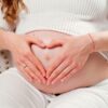 Imagem de uma mulher grávida, sentada, com a barriga descoberta e fazendo um coração nela com as mãos