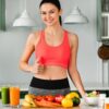 Mulher branca com top na frente de uma mesa com frutas e copos de suco.