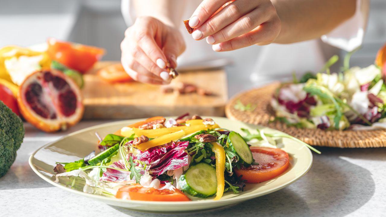 Foto mostra um prato de salada em cima da mesa enquanto uma moça joga sal em saquinho.