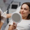 Foto mostra uma mulher sentada numa mesa médica com um espelho ao lado de uma médica.