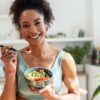 Mulher negra comendo frutas em uma cumbuca e olhando para a câmera sorrindo.