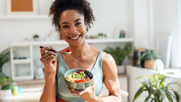 Mulher negra comendo frutas em uma cumbuca e olhando para a câmera sorrindo.