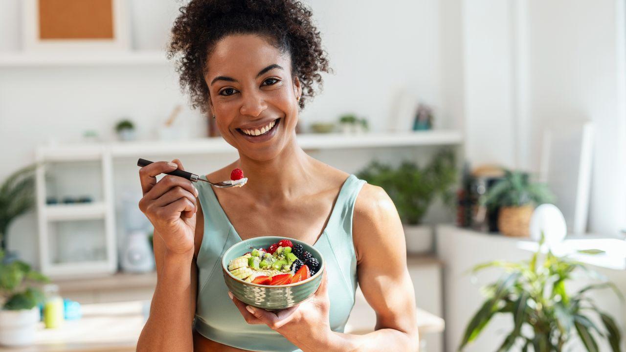Mulher negra comendo frutas em uma cumbuca e olhando para a câmera sorrindo.