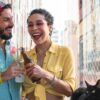 Mulher e homem com roupas coloridas brindando uma cerveja.