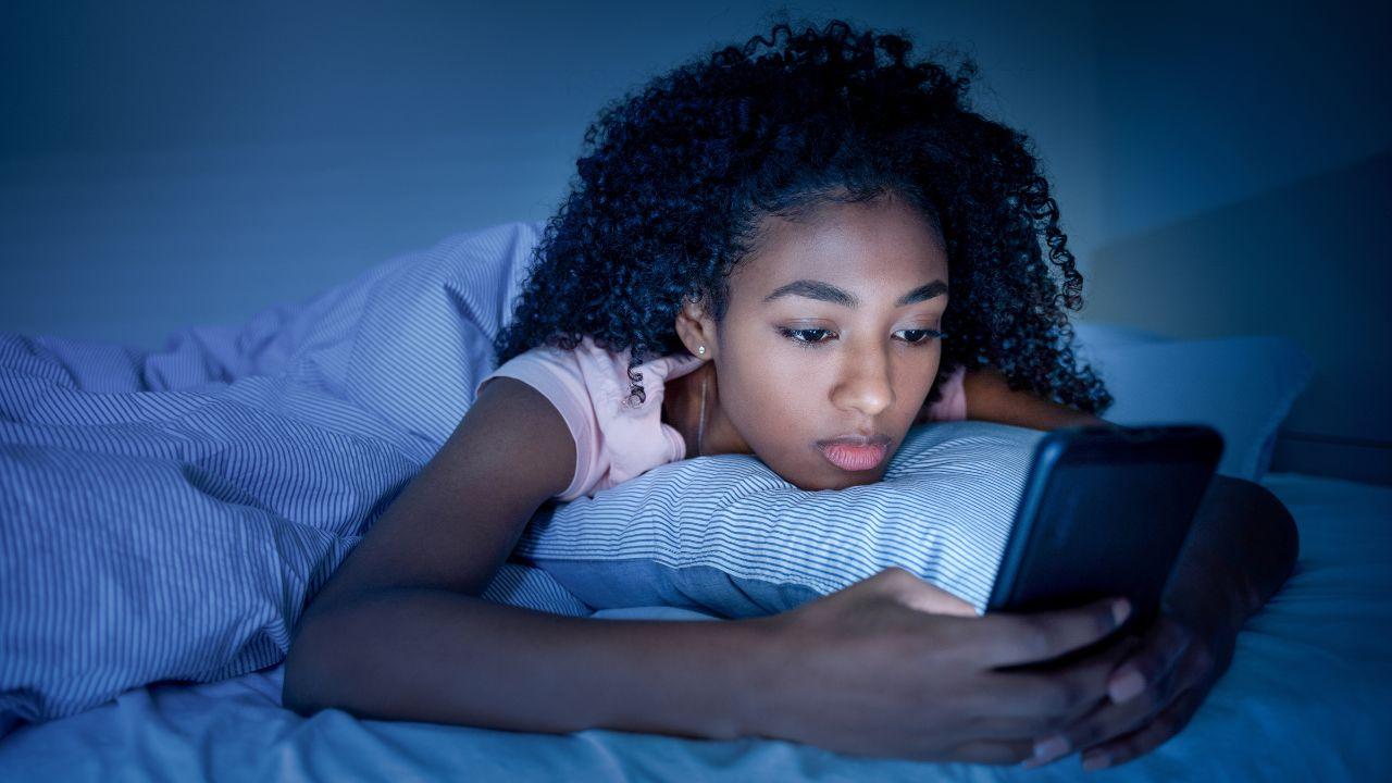 Mulher negra mexendo no celular deitada na cama a noite.