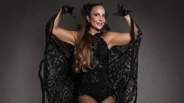 A foto mostra Ivete pronta pra o show de Porto Seguro. Ela veste body preto brilhante com transparências e detalhes rendados, meia-calça arrastão e sandálias pretas de salto alto. Sobre os ombros, usa uma capa ampla de renda preta que se abre como asas. O cabelo está longo, com adorno preto na lateral da cabeça.
