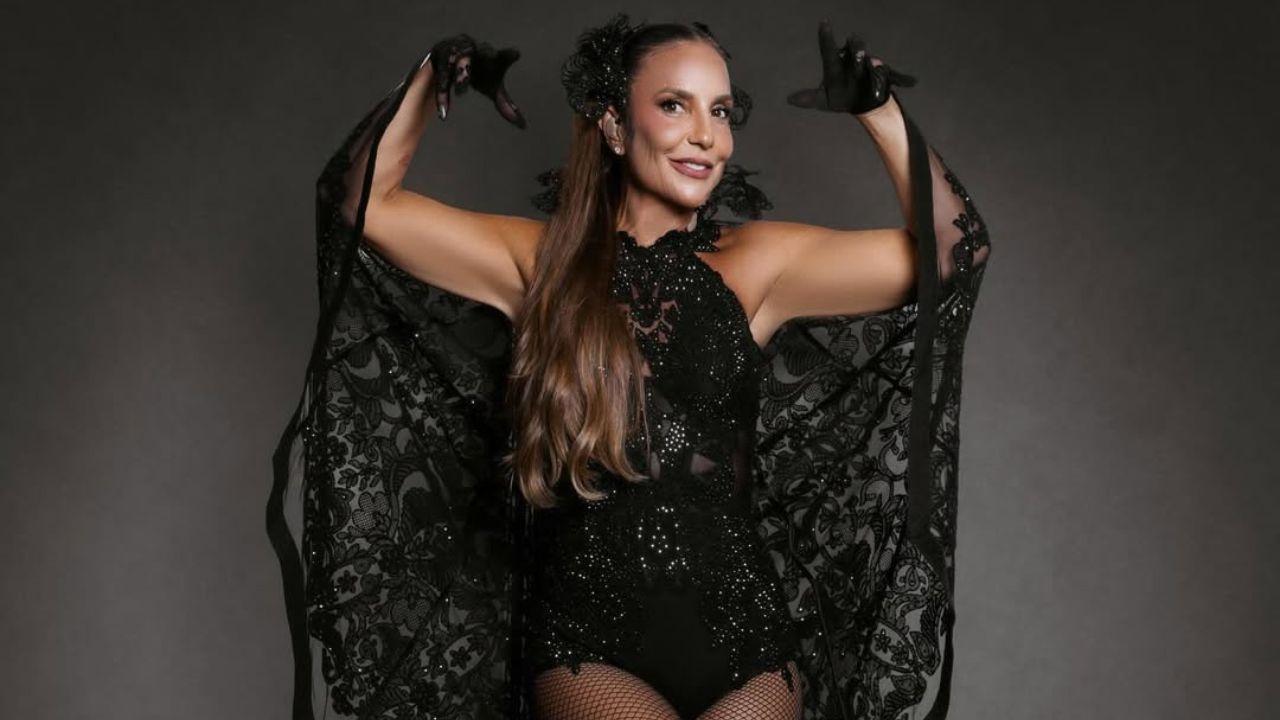 A foto mostra Ivete pronta pra o show de Porto Seguro. Ela veste body preto brilhante com transparências e detalhes rendados, meia-calça arrastão e sandálias pretas de salto alto. Sobre os ombros, usa uma capa ampla de renda preta que se abre como asas. O cabelo está longo, com adorno preto na lateral da cabeça.