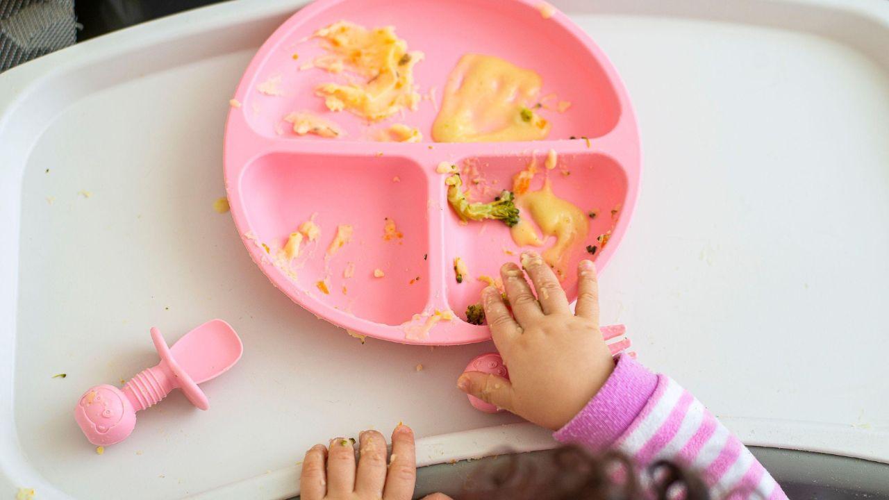 Imagem de uma criança pegando comida de seu prato com divisórias com a mão
