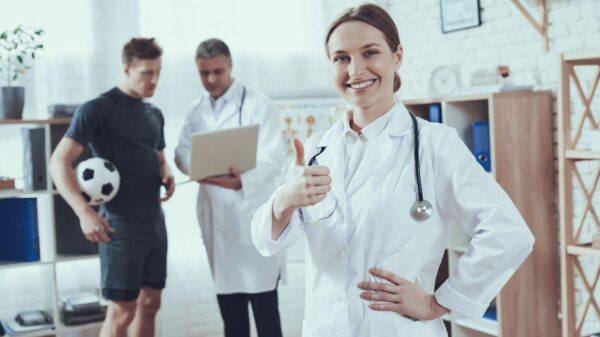 Imagem de uma médica fazendo joia com o dedão e um médico conversando com um atleta no fundo