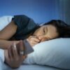 Foto mostra mulher deitada na cama no escuro e mexendo no celular.