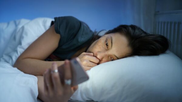 Foto mostra mulher deitada na cama no escuro e mexendo no celular.