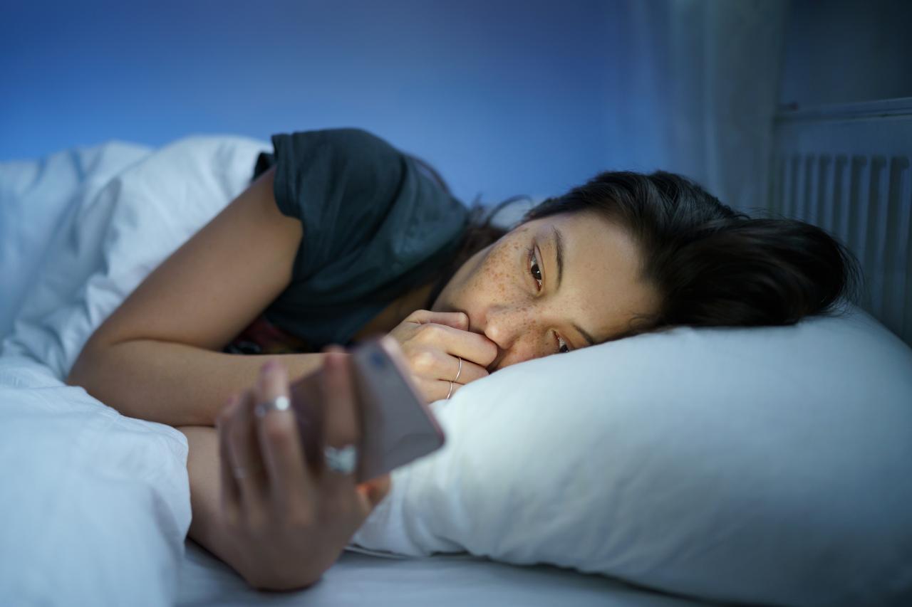 Foto mostra mulher deitada na cama no escuro e mexendo no celular.