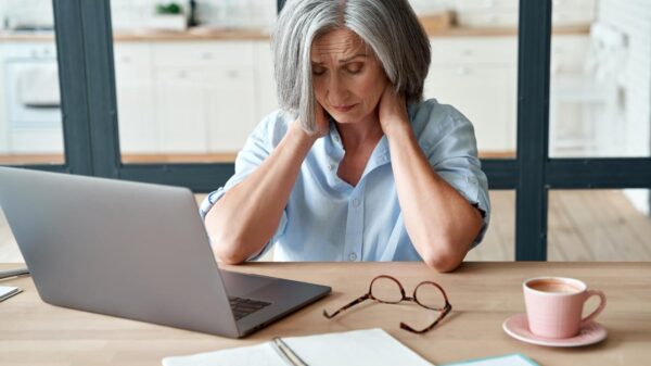 Mulher de 50 anos, de cabelo branco, sentada no computador com rosto preocupado.