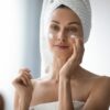 Mulher com toalha no corpo e na cabeça passando creme no rosto.