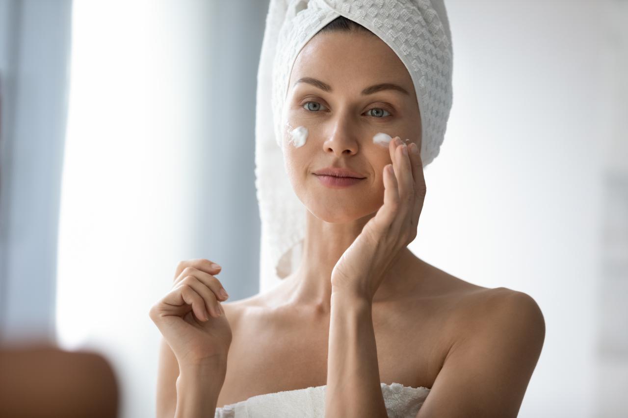 Mulher com toalha no corpo e na cabeça passando creme no rosto.