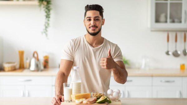 Homem na cozinha, na frente da mesa com comidas saudáveis. Ele faz o sinal de joinha.