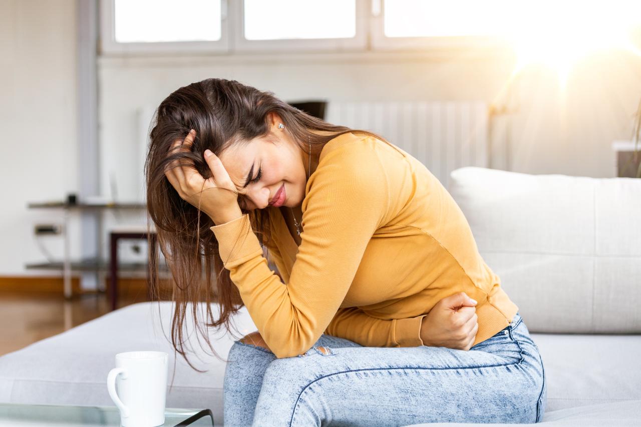 Mulher sentada no sofá com a mão na barriga apresentando desconforto abdominal.