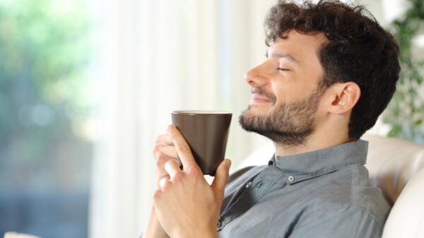 Homem branco, com barba, cabelo castanho, sentado no sofá, segurando uma xícara de café.
