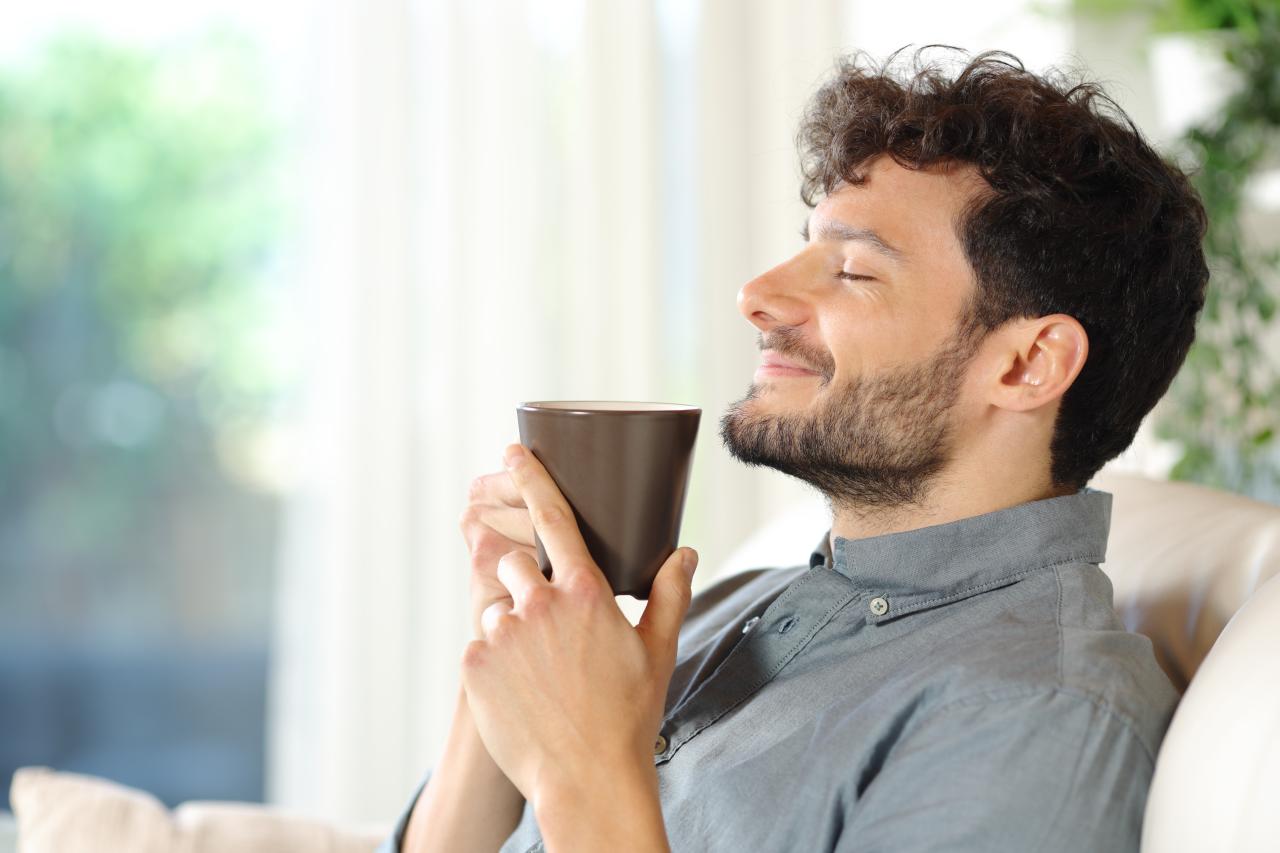 Homem branco, com barba, cabelo castanho, sentado no sofá, segurando uma xícara de café.