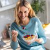 Mulher comendo um potinho de iogurte com frutas em pé e sorrindo.