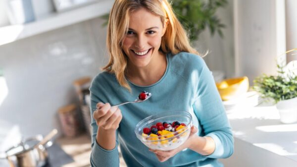 Mulher comendo um potinho de iogurte com frutas em pé e sorrindo.