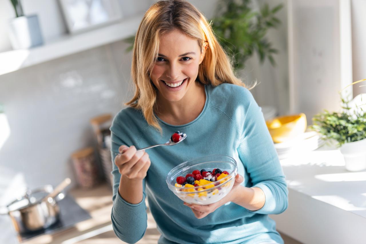 Mulher comendo um potinho de iogurte com frutas em pé e sorrindo.