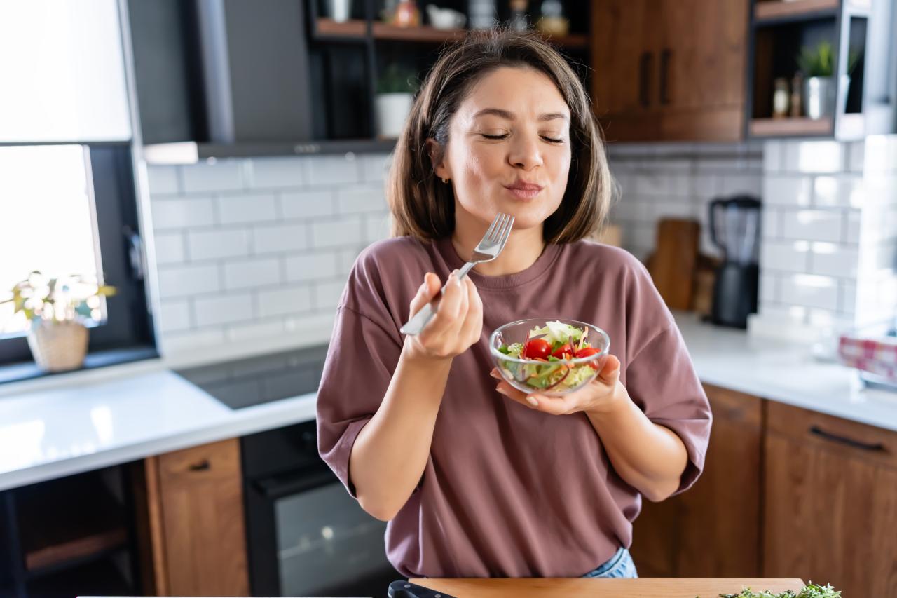 Mulher branca, em pé, cabelo castanho, comendo comida saudável em um pote de vidro.