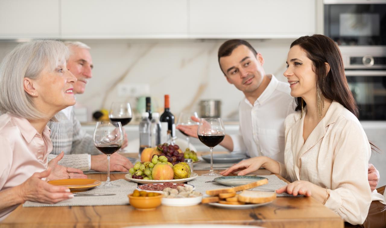 Um casal jovem sentados numa mesa enquanto comem em frente a um casal de idosos que são pais de algum deles.