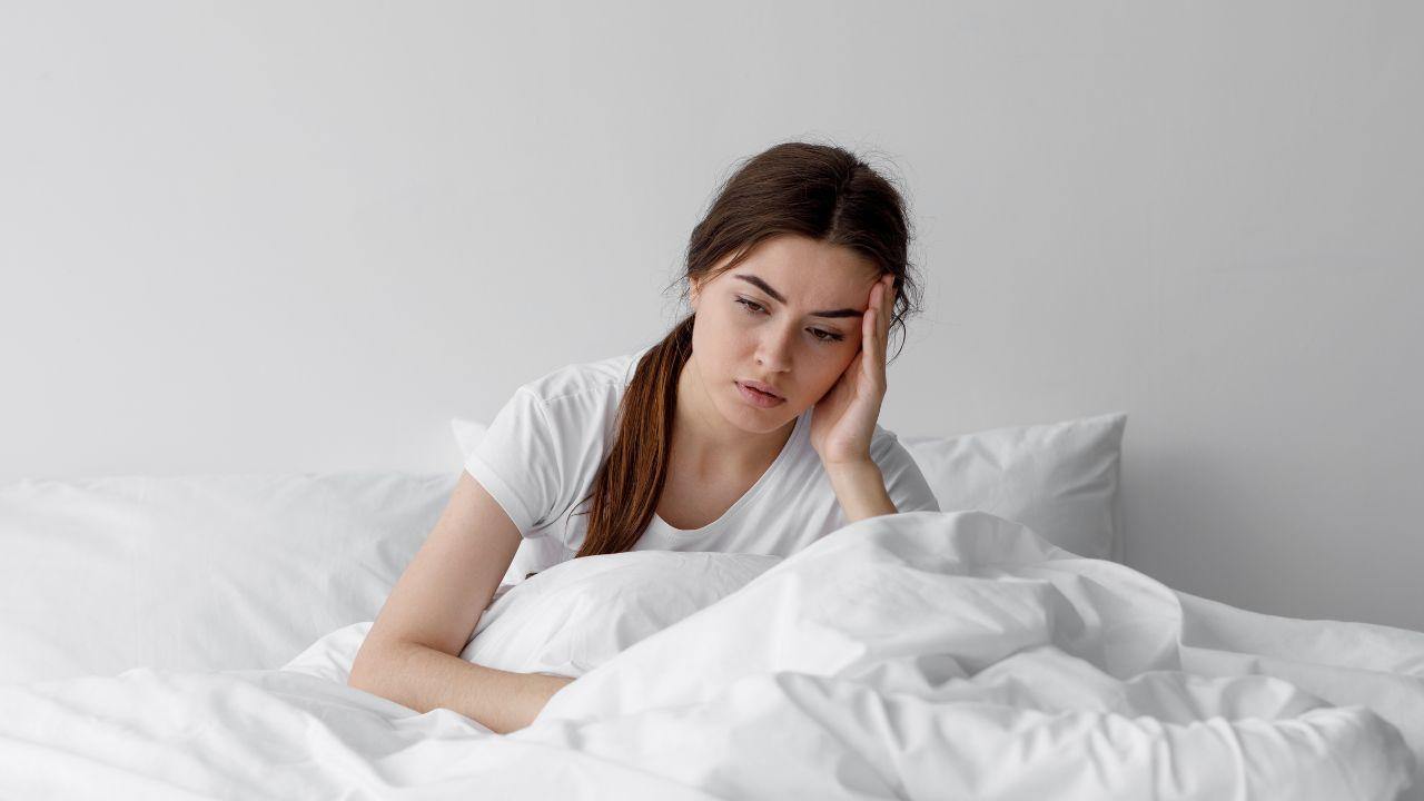 Imagem de uma mulher sentada na cama, com cobertas brancas cobrindo suas pernas, e a mão na cabeça